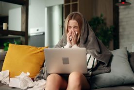 A white female sits on a modern sofa blowing her nose, she is wrapped in a blanket, surrounded by tissues and has a laptop on her knees.