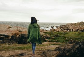 A woman walks along a grass path near to the ocean, wearing a coat on a grey day.