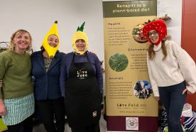 Four women stand in front a tall banner wearing hats in the shapes of a banana, pineapple and cherry.