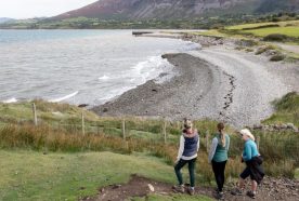 Three women pause on a low cliff looking out over a calm bay with brown water and distant hills.