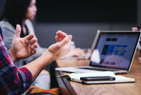 A close up of a man's hands next to a laptop with a person in the background discussing issues around a wooden table.